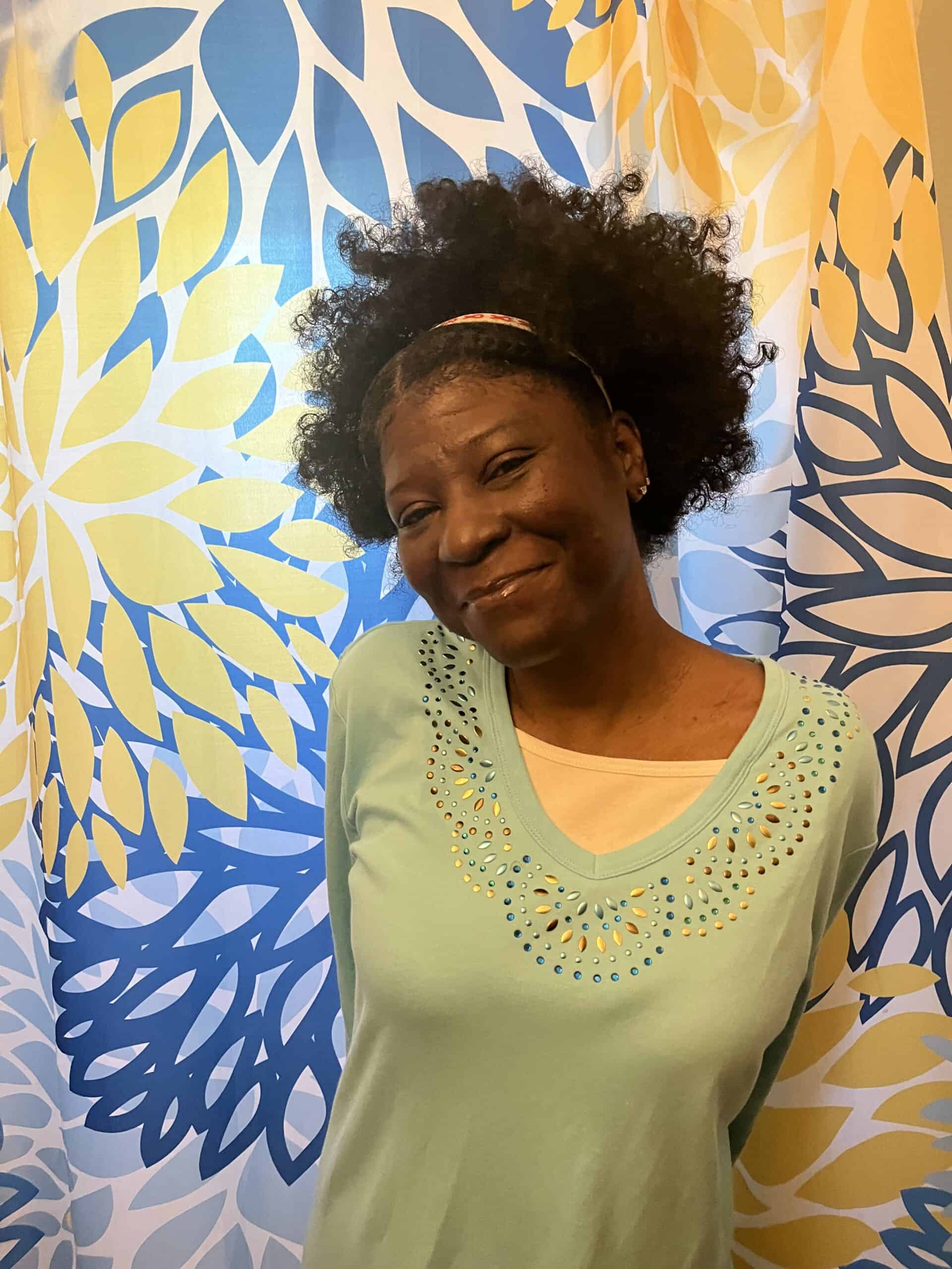 A woman rich in melanin smiles in front of a colorful wall full of flowers. She is wearing a green sweater layered over a yellow shirt, and a yellow headband.