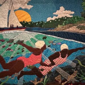 This quilt depicts a pair of black women swimming in the shore, a sailboat behind them. The setting sun behind the boat.