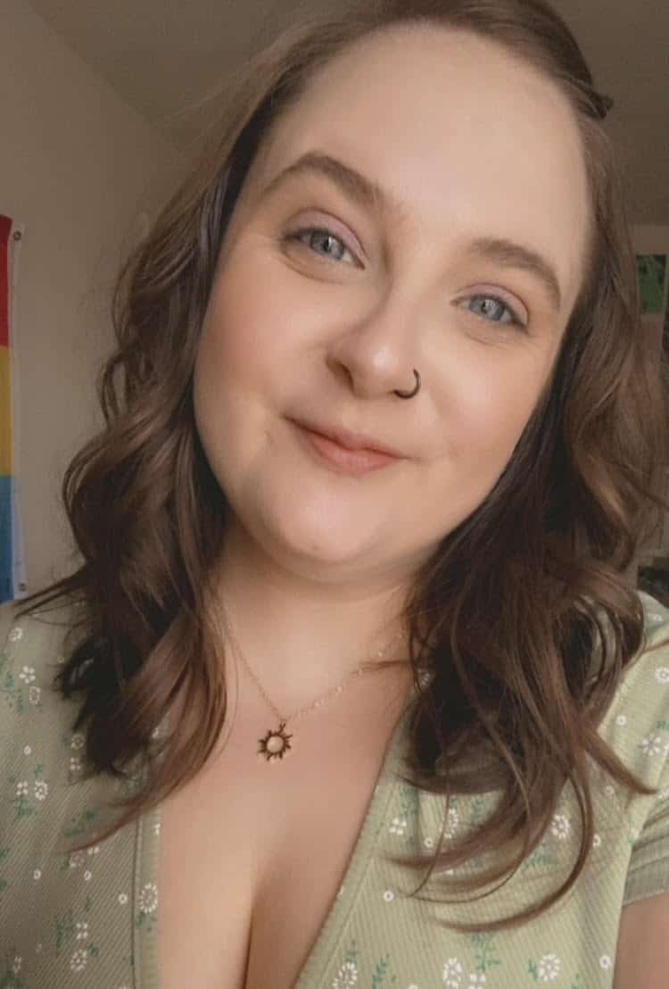 A person with shoulder length auburn hair smiles at the camera. They are wearing a green top and necklace.