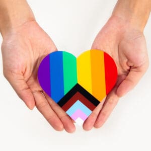 Two hands hold a heart decorated like the pride progress flag.