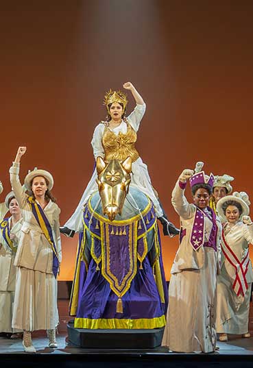 A picture from the musical "Suffs" it shows women in Victorian outfits with suffragette sashes. One is riding a metal horse.