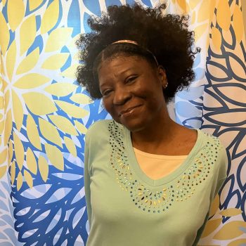 A woman rich in melanin smiles in front of a colorful wall full of flowers. She is wearing a green sweater layered over a yellow shirt, and a yellow headband.