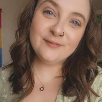 A person with shoulder length auburn hair smiles at the camera. They are wearing a green top and necklace.