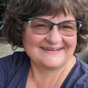 A fair-skinned woman with short, curly hair and cat-eyed glasses wears a blue blouse and smiles at the camera.