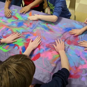 Children put their hands on a tie-die table cloth.
