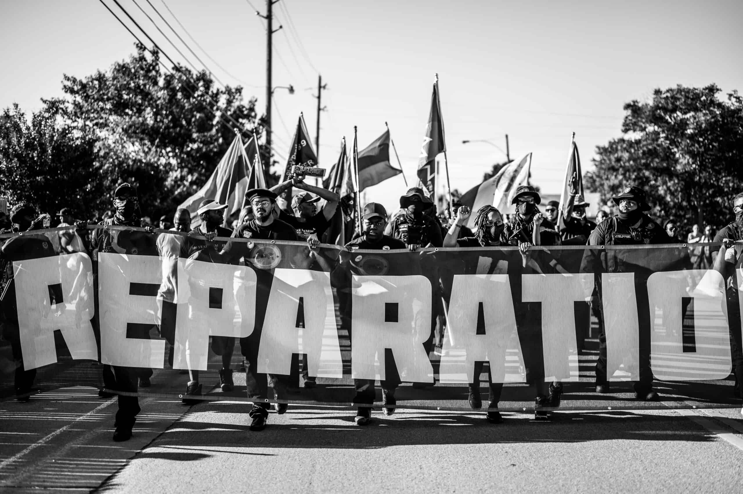 A black and white photos show markers carrying a sign that say "Reparations."
