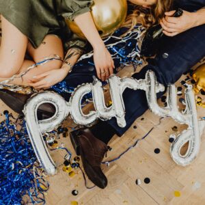 Two white people sit on the floor among party decorations. On their laps are a set of balloons that spell out "Party."