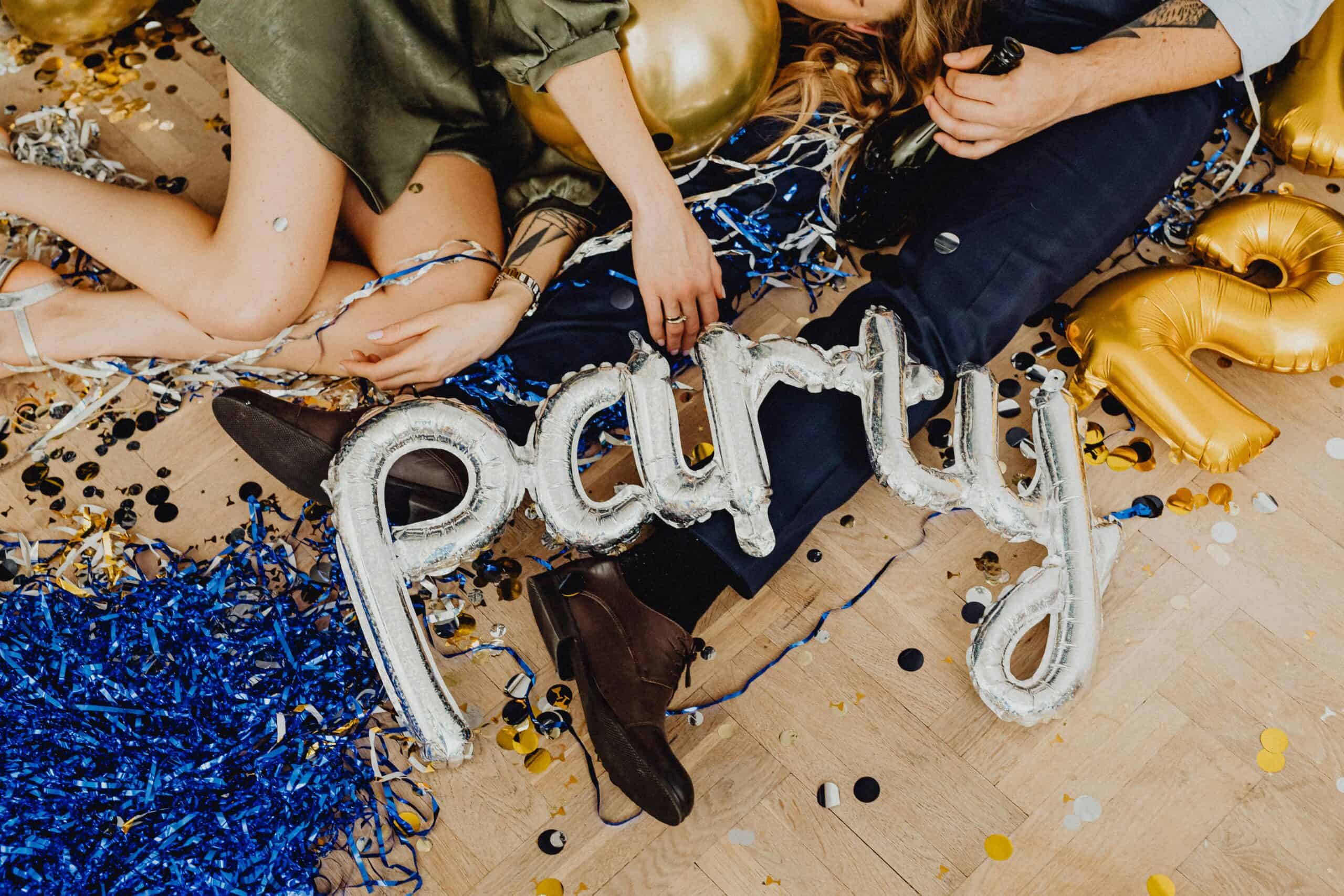 Two white people sit on the floor among party decorations. On their laps are a set of balloons that spell out "Party."