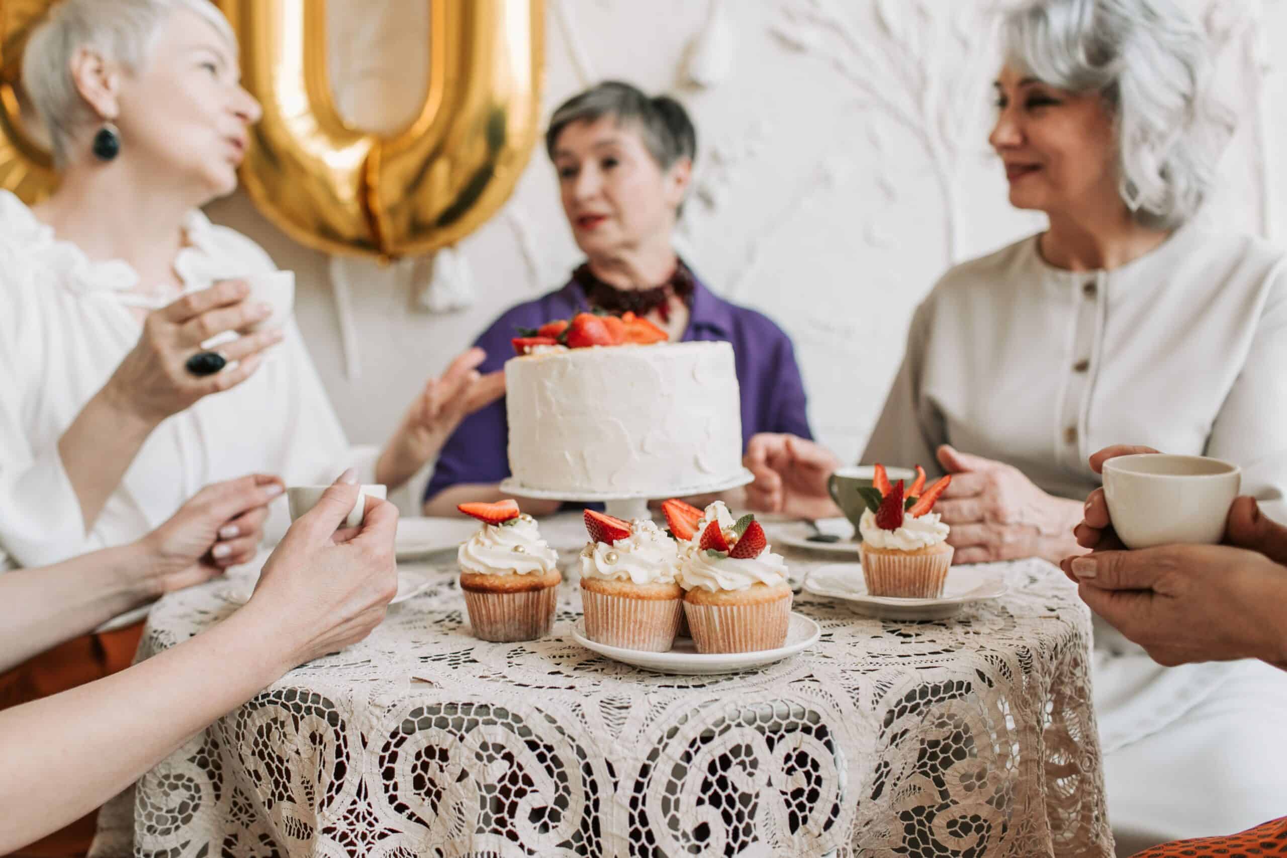 Women with grey hair gather around a table with cake.