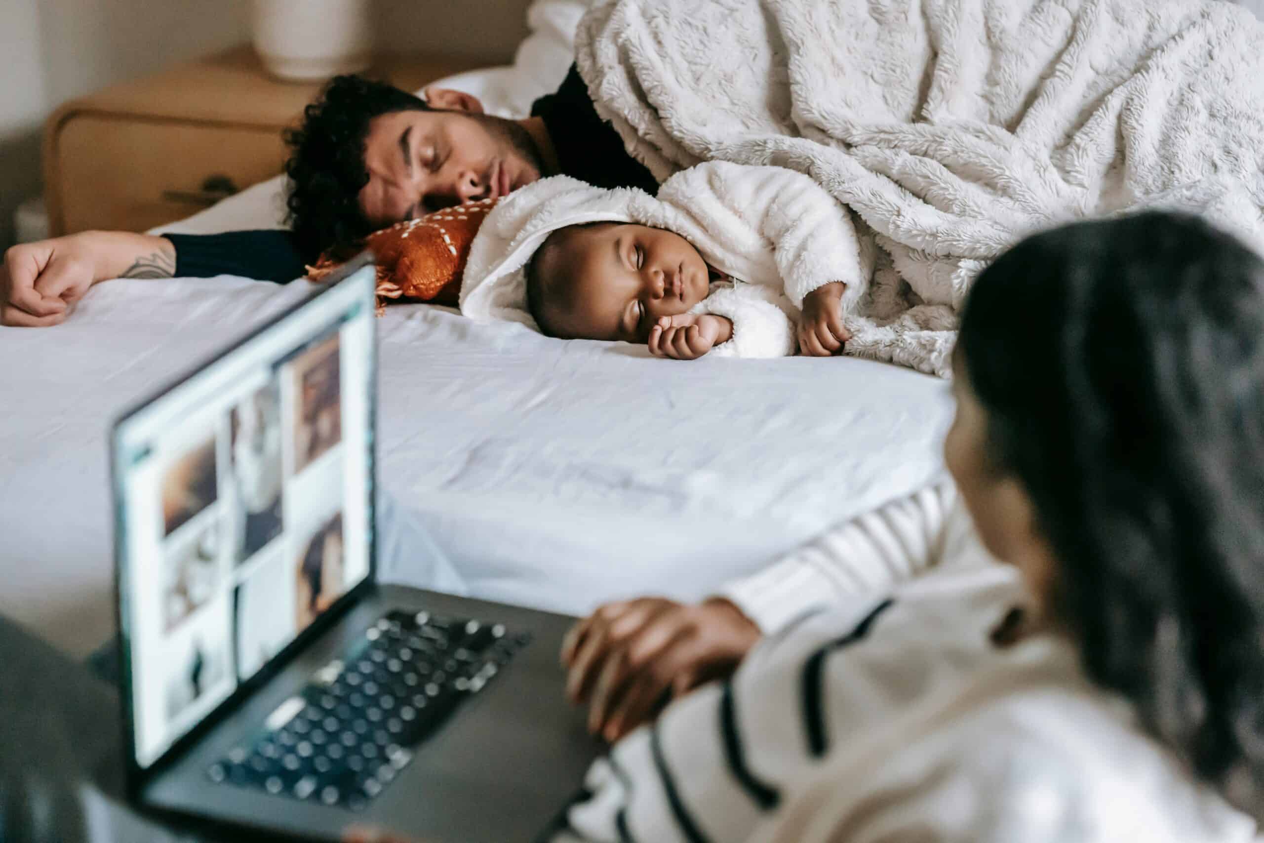 A dad and his baby sleep under cozy blankets while the child's mother watches them adoringly.