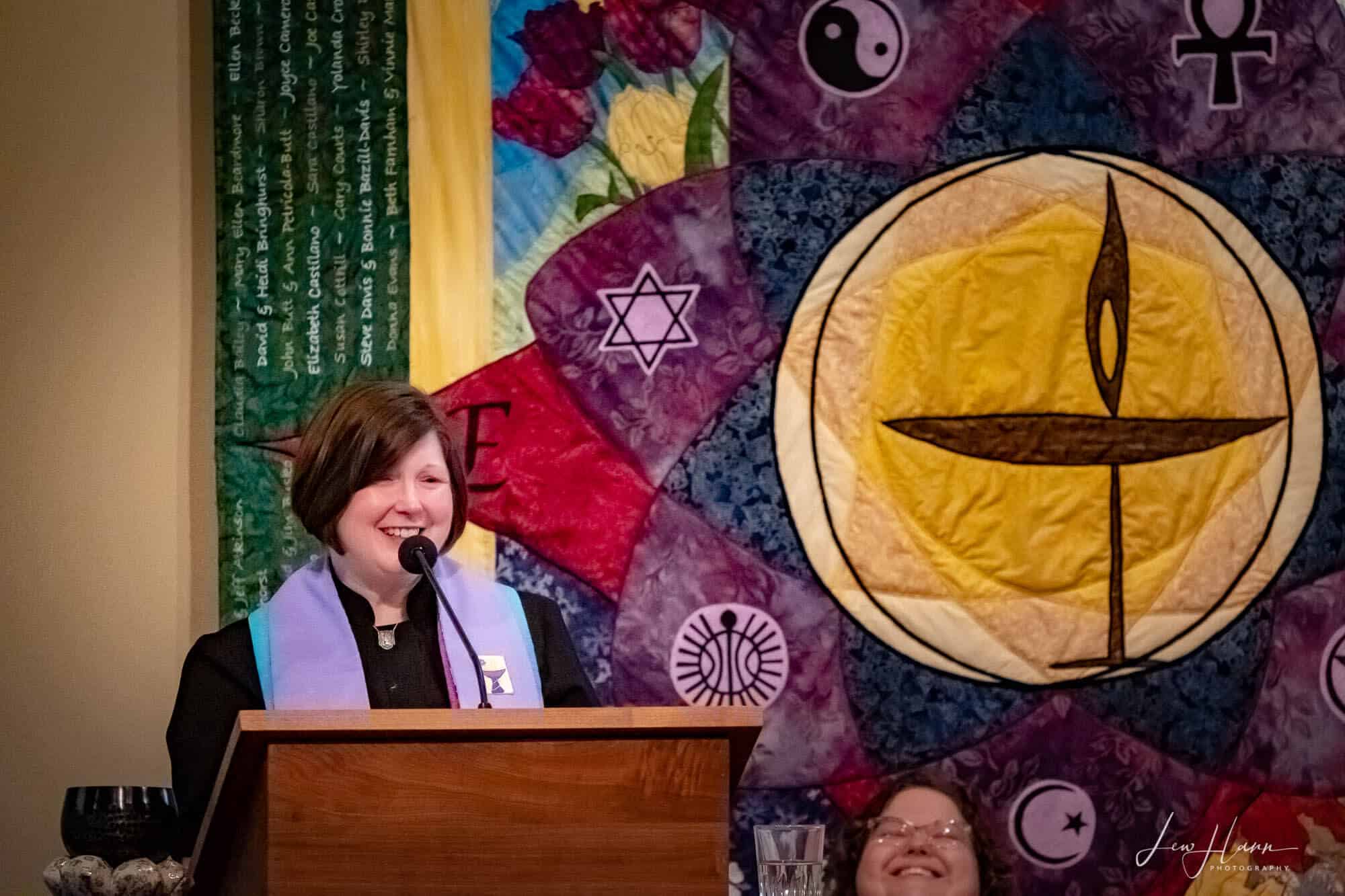 Rev. Kellie stands at the pulpit, in front of our congregation's tapestry.